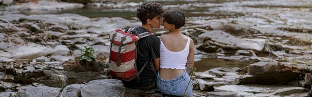 Back view of couple of hikers sitting on bank of rocky river and kissing. Hiking in mountains. Rear. - Фото, зображення