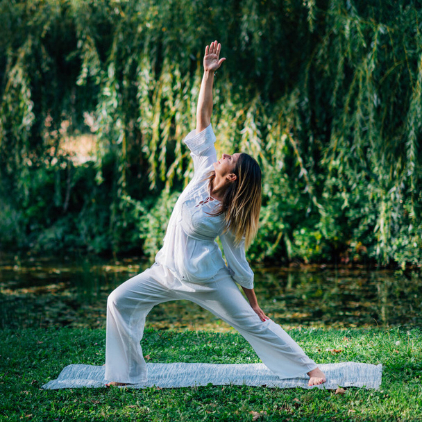 Yoga in nature, young woman in white, peaceful warrior pose, soft green nature background. - 写真・画像