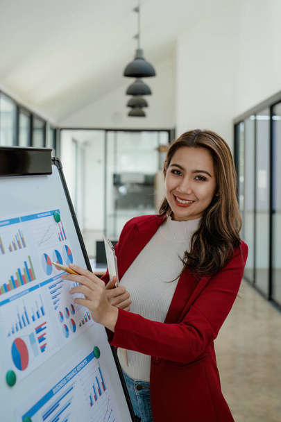 portrait of Beautiful young asian businesswoman presents business profits to colleagues at meeting, explaining business turn over on flipchart to coworkers in office with using tablet. - 写真・画像