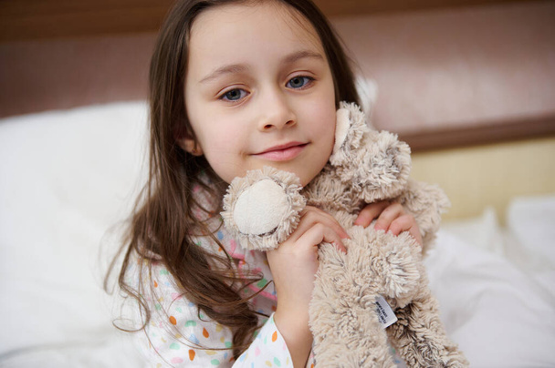 Close-up portret van een heerlijk schattig klein meisje met mooie lange horen, het dragen van een pyjama, zachtjes knuffelen van haar pluche speelgoed en glimlachend kijken naar de camera terwijl wakker in de ochtend op het bed - Foto, afbeelding