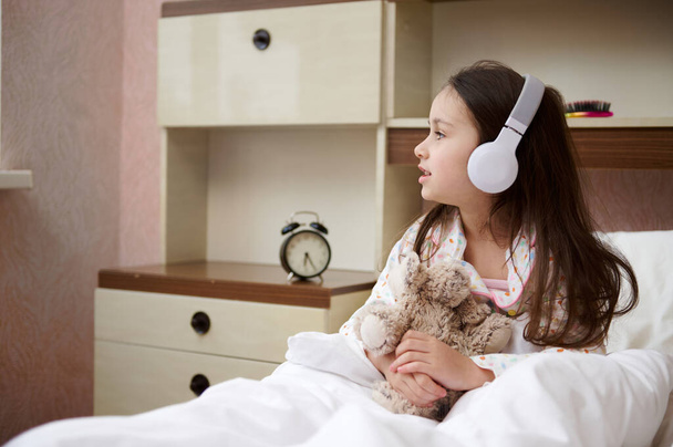 Happy European elementary age child girl in glamour pajama, sitting with a plush toy sheep in her hands on bed when waking up, listening to music on wireless headphones and looking aside at the window - Foto, afbeelding