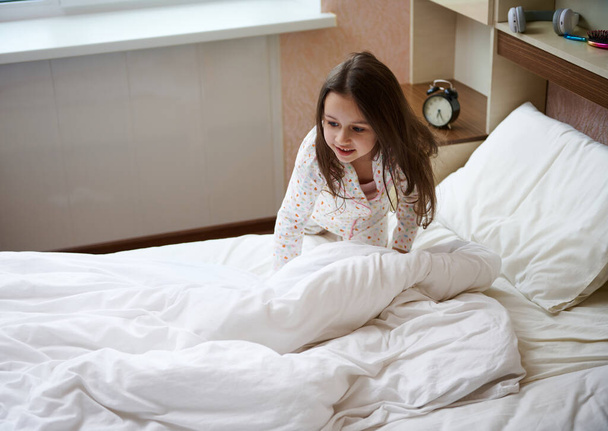 Ihana onnellinen valkoihoinen lapsi tyttö tyylikkäässä valkoisessa pyjamassa, jossa on värikkäitä pisteitä, herääminen onnellisella tuulella, hymyillen ja taittamalla sänkynsä ja puhdistamalla makuuhuoneensa aurinkoisena aamuna. - Valokuva, kuva
