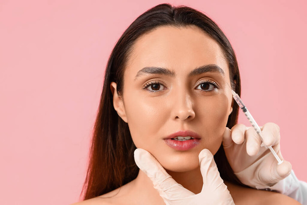Young woman receiving filler injection in face against pink background - 写真・画像