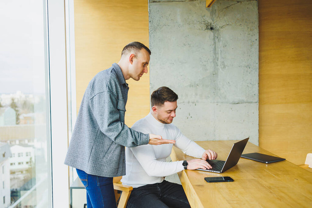 Zwei männliche Kollegen bei der Arbeit mit einem Laptop im Büro. Zwei zielorientierte Unternehmer arbeiten in einem modernen Arbeitsraum zusammen. Zwei junge Geschäftsleute arbeiten in einem hellen Büro - Foto, Bild