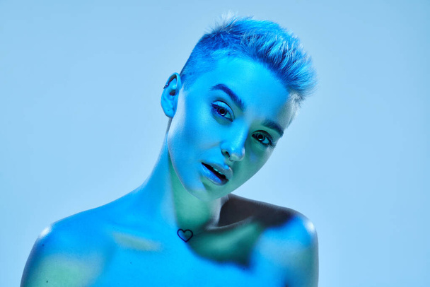 Beautiful young girl with short blonde hair posing with bare shoulders against blue studio background in neon light. Cyberpunk style. Technology, modern fashion, digital art, virtual reality concept - Photo, Image