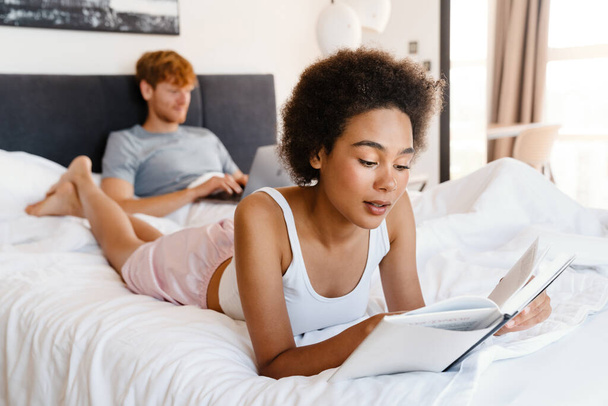 Young beautiful african curly woman reading book lying with her boyfriend who working with laptop on bed at home - Fotó, kép