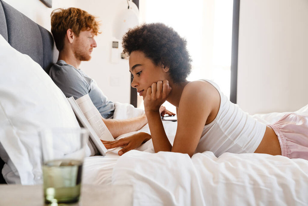 Young beautiful african curly woman propping her head and reading book lying with her boyfriend who working with laptop on bed at home - Фото, изображение