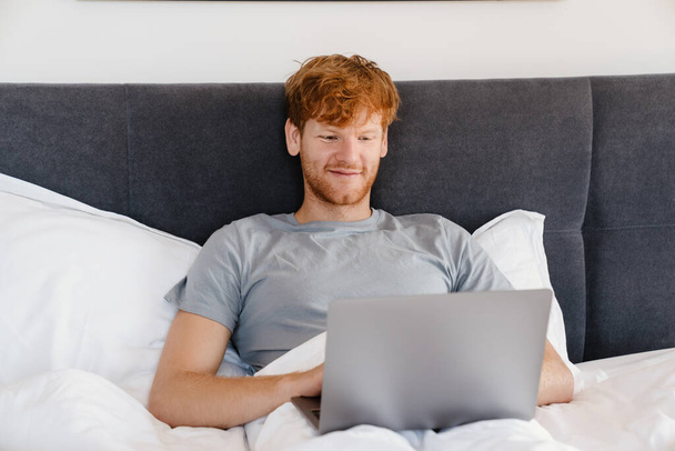 Young handsome smiling redhead man in gray t-shirt working with laptop, while liying on bed at home - Фото, зображення