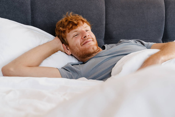 Νεαρός όμορφος χαμογελαστός κοκκινομάλλης με γκρι μπλουζάκι με το χέρι πίσω από το κεφάλι του κοιτάζοντας στην άκρη ξαπλωμένος στο κρεβάτι στο σπίτι - Φωτογραφία, εικόνα