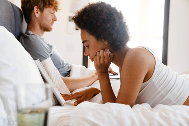 Young beautiful african curly woman propping her head and reading book lying with her boyfriend who working with laptop on bed at home - Φωτογραφία, εικόνα