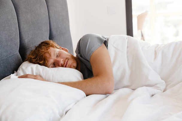 Portrait of young handsome sleeping redhead man in gray t-shirt on bed at home - Fotografie, Obrázek