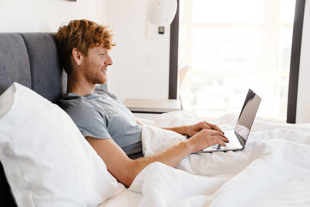 Young handsome smiling redhead man in gray t-shirt working with laptop, while liying on bed at home - Fotoğraf, Görsel