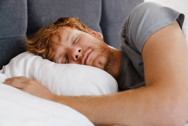 Portrait of young handsome sleeping redhead man in gray t-shirt on bed at home - Photo, image