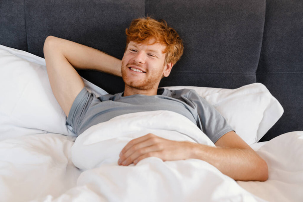 Young handsome smiling redhead man in gray t-shirt with hand behind his head looking aside lying on bed at home - Foto, Imagem