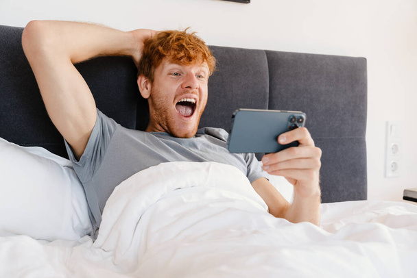 Young handsome enthusiastic redhead man in gray t-shirt holding his head and looking at his phone, while liying on bed at home - Φωτογραφία, εικόνα