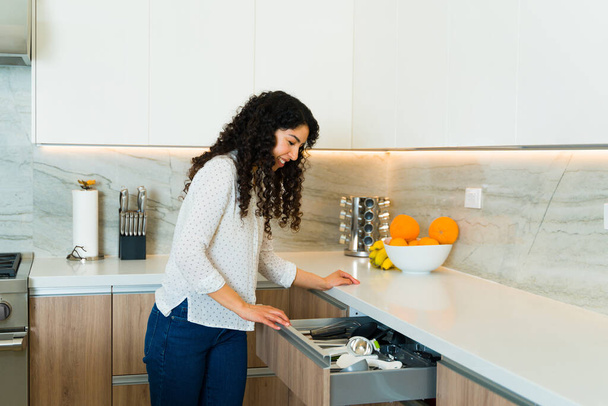 Joyeux belle femme se préparer à cuisiner et à la recherche d'une cuillère sur le tiroir en granit dans sa cuisine de luxe - Photo, image