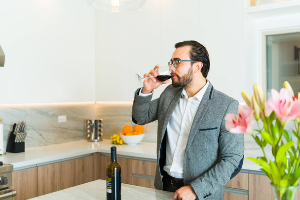 Hombre caucásico en sus 30 años bebiendo una copa de vino mientras espera en su cocina de lujo para una cita formal en casa - Foto, imagen