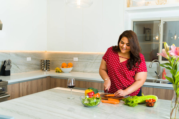 Latynoska otyła kobieta uśmiecha się podczas krojenia warzyw do gotowania obiadu w swojej granitowej, luksusowej kuchni - Zdjęcie, obraz