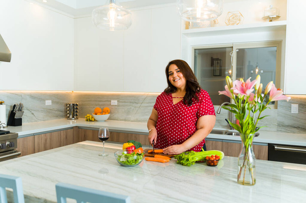 Latin big woman smiling while cutting vegetables to make a healthy salad on the white quartz kitchen island - 写真・画像