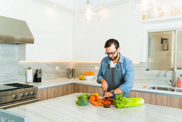 Όμορφος καυκάσιος άνδρας χαμογελά φορώντας μια ποδιά ενώ είναι έτοιμος να μαγειρέψει ένα υγιεινό γεύμα σε μια όμορφη πολυτελή κουζίνα - Φωτογραφία, εικόνα