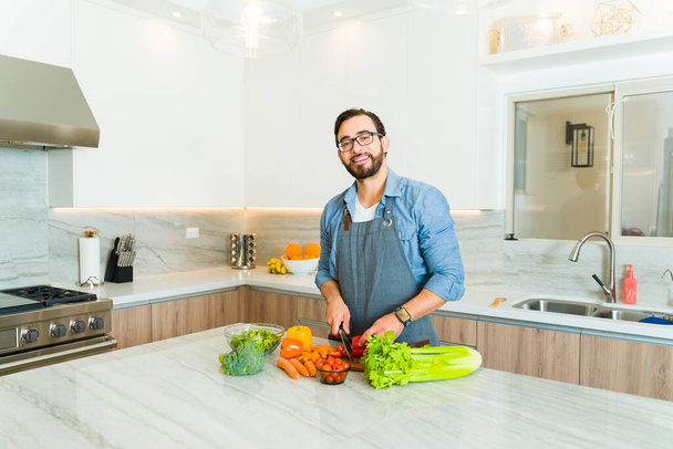 Viehättävän 30-vuotiaan miehen muotokuva leikkelemässä vihanneksia ja tekemässä terveellistä illallista graniittisella keittiösaarella. - Valokuva, kuva