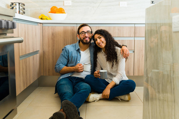 Alegre hombre caucásico y mujer hispana abrazándose mientras sonríen tomando café juntos en el piso de la cocina - Foto, imagen
