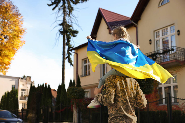Askeri üniformalı asker, kızını omuzlarında taşıyor ve Ukrayna bayrağı açık havada, mesaj için uzayda. Aile toplantısı - Fotoğraf, Görsel