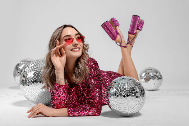 Beautiful woman in sunglasses and sequin dress among disco balls on white background - Fotografie, Obrázek