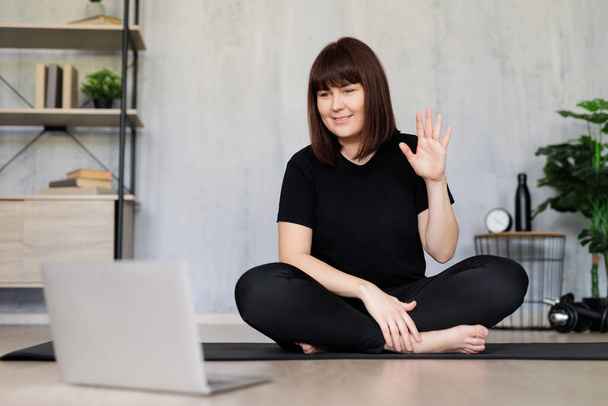 joven hermosa mujer sentada en estera de yoga y haciendo yoga con tutorial en línea en el ordenador portátil - Foto, imagen