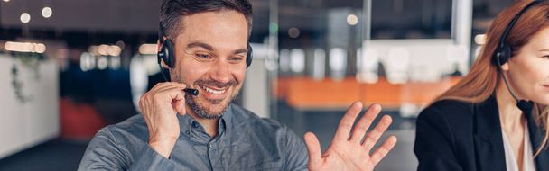 Hymyilevä liikemies kuulokkeissa puhuu verkossa asiakkaan kanssa kollegan taustalla - Valokuva, kuva