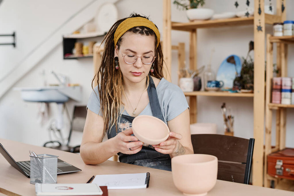 Jonge vrouw die handgemaakt aardewerk boven tafel houdt terwijl ze in de studio of werkplaats zit en de kwaliteit van de kleikom controleert - Foto, afbeelding