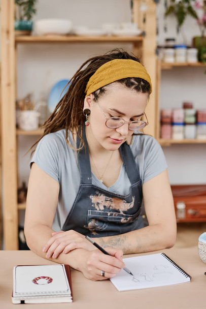Jeune potier femme créative faisant un croquis de produits en faïence dans un bloc-notes assis à table dans un petit magasin ou un studio - Photo, image