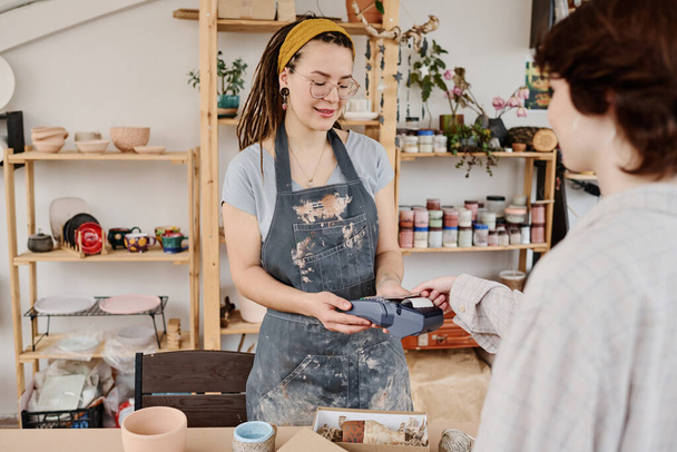 Молода жінка-власник магазину невеликого глиняного посуду з платіжним терміналом, що продає предмети ручної роботи клієнту, який платить за покупку кредитною карткою
 - Фото, зображення