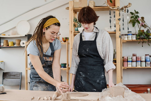 Молода жінка-майстер консультує свого учня про створення предметів глиняного посуду, вказуючи при цьому на плоский шматок глини
 - Фото, зображення