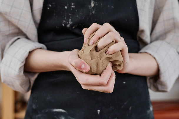 Ruce mladé kreativní ženy v pracovním oděvu drží kus měkké hlíny a hnětení ji při přípravě na výrobu hliněných výrobků - Fotografie, Obrázek