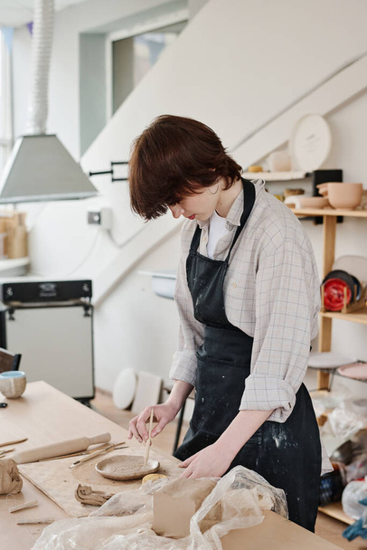 Mujer joven en ornamento de corte de ropa de trabajo en placa de arcilla cruda mientras se inclina sobre la mesa con suministros para la creación de loza - Foto, Imagen
