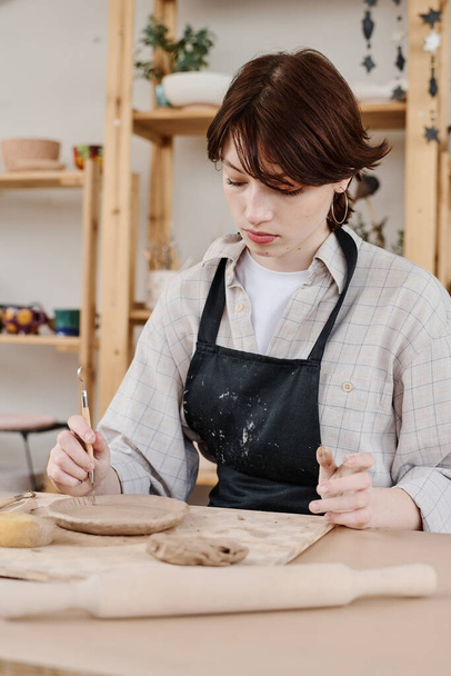 Bruneta žena v zástěře sedí u pracoviště a používá dřevěný ruční nástroj pro řezání ozdoby na povrchu surového jílu - Fotografie, Obrázek