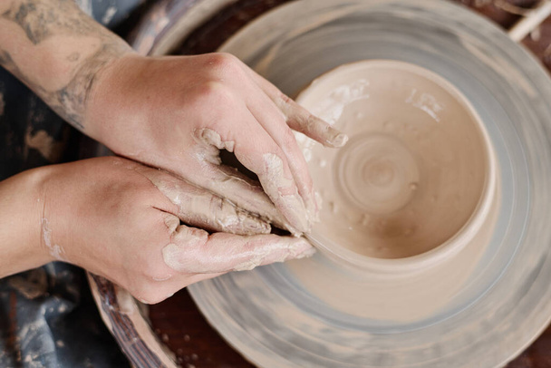 Visão geral das mãos de jovem artesão formando forma de jarro de barro ou panela girando na roda de cerâmica, enquanto cerâmica creatina - Foto, Imagem