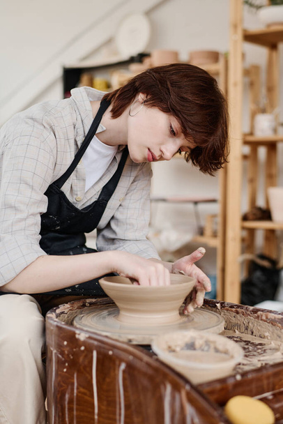 Young female pitcher bending over rotating pottery wheel during work over new clay item while sitting in her workshop or studio - Foto, Imagem
