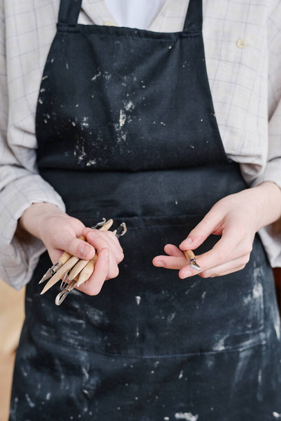 Mãos de jovem mulher artesã em avental preto segurando grupo de ferramentas manuais de madeira necessárias para cortar ornamentos na superfície de itens de argila - Foto, Imagem