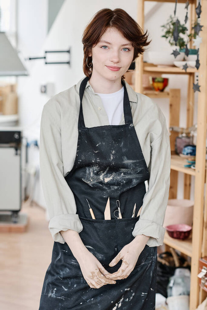 Young creative female pitcher in workwear standing in front of camera against interior of workshop or studio and wooden shelves - Photo, Image