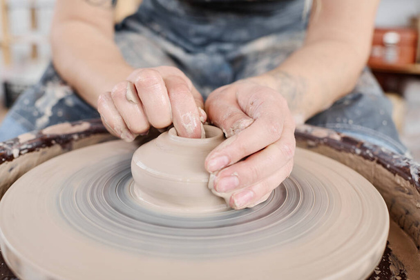 Руки молодой женщины-гончара на сырой глине на вращающемся гончарном круге во время создания новой глиняной посуды для продажи в мастерской - Фото, изображение