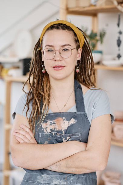 Νεαρή γυναίκα υπάλληλος με αυτοπεποίθηση ή ιδιοκτήτρια ενός μικρού μαγαζιού με πήλινα σκεύη που σας κοιτάζει ενώ στέκεται απέναντι στην έκθεση με χειροποίητα πήλινα αντικείμενα - Φωτογραφία, εικόνα