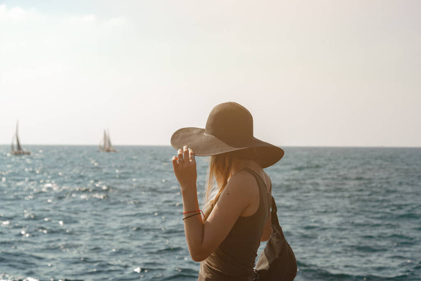 Musta hattuinen tyttö katsoo kohti merta. Kaksi purjeveneitä purjehtii horisontissa. Malli trimmaa hatun aurinkoisella säällä meren rannalla. Kehys on valaistu aurinko - Valokuva, kuva