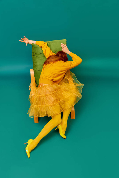 Creative portrait of redhead girl with long straight hair posing on armchair, sitting in strange poses over cyan color background. Impersonal emotions, body language. Fashion, beauty, mental health - Photo, Image