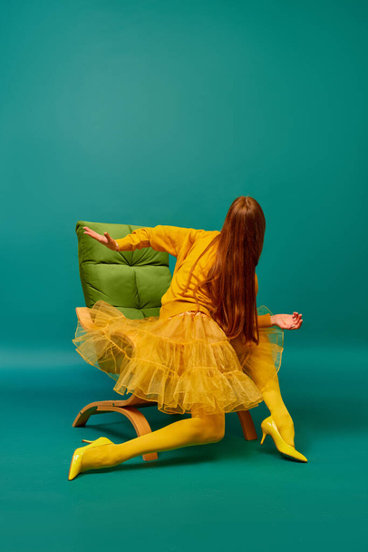 Creative portrait of redhead girl with long straight hair posing on armchair, sitting in strange poses over cyan color background. Impersonal emotions, body language. Fashion, beauty, mental health - Fotografie, Obrázek
