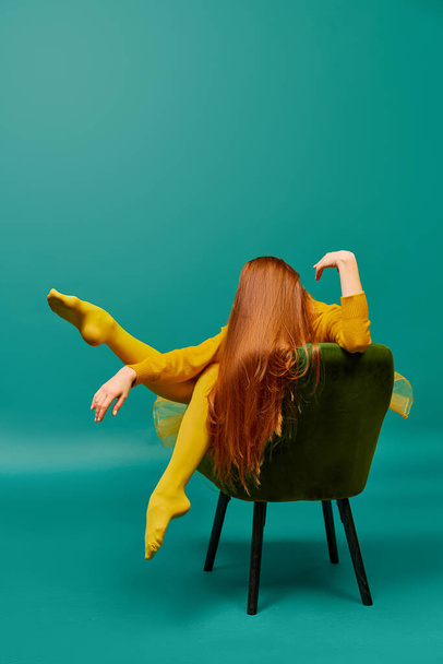 Creative portrait of redhead girl with long straight hair posing on armchair, sitting in strange poses over cyan color background. Impersonal emotions, body language. Fashion, beauty, mental health - Photo, Image