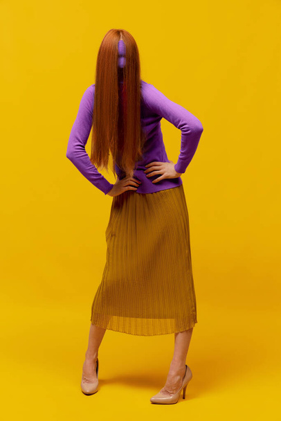 Outojen ihmisten käsite. Kasvoton muotokuva hoikka nainen pitkät suorat punaiset hiukset yllään retro muoti asu toiminnassa yli keltainen studio tausta. persoonattomat tunteet, kehon kieli - Valokuva, kuva