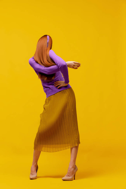 Παράξενος χορός. Πρόσωπο πορτρέτο της λεπτής γυναίκας με μακριά ίσια κόκκινα μαλλιά φορώντας ρετρό ρούχα μόδας σε δράση πάνω από κίτρινο φόντο στούντιο. Απροσωπικά συναισθήματα, γλώσσα του σώματος - Φωτογραφία, εικόνα
