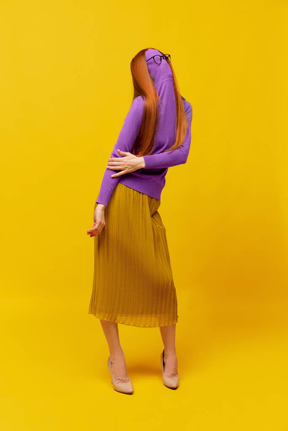 Be different. Faceless portrait of slim woman with long straight red hair wearing retro fashion outfit in action over yellow studio background. Impersonal emotions, body language - Photo, image